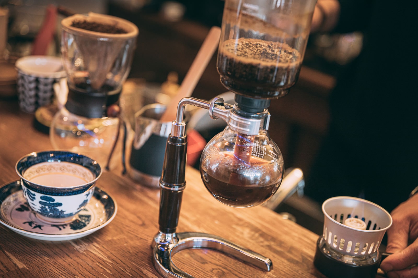 An empty coffee cup sits next to a coffee siphon coffee maker brewing coffee on a counter